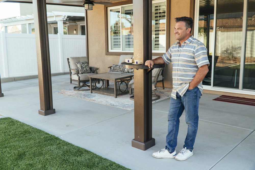 Smiling man leaning against an Alumatable mounted on his patio cover post with two drinks also resting on the table